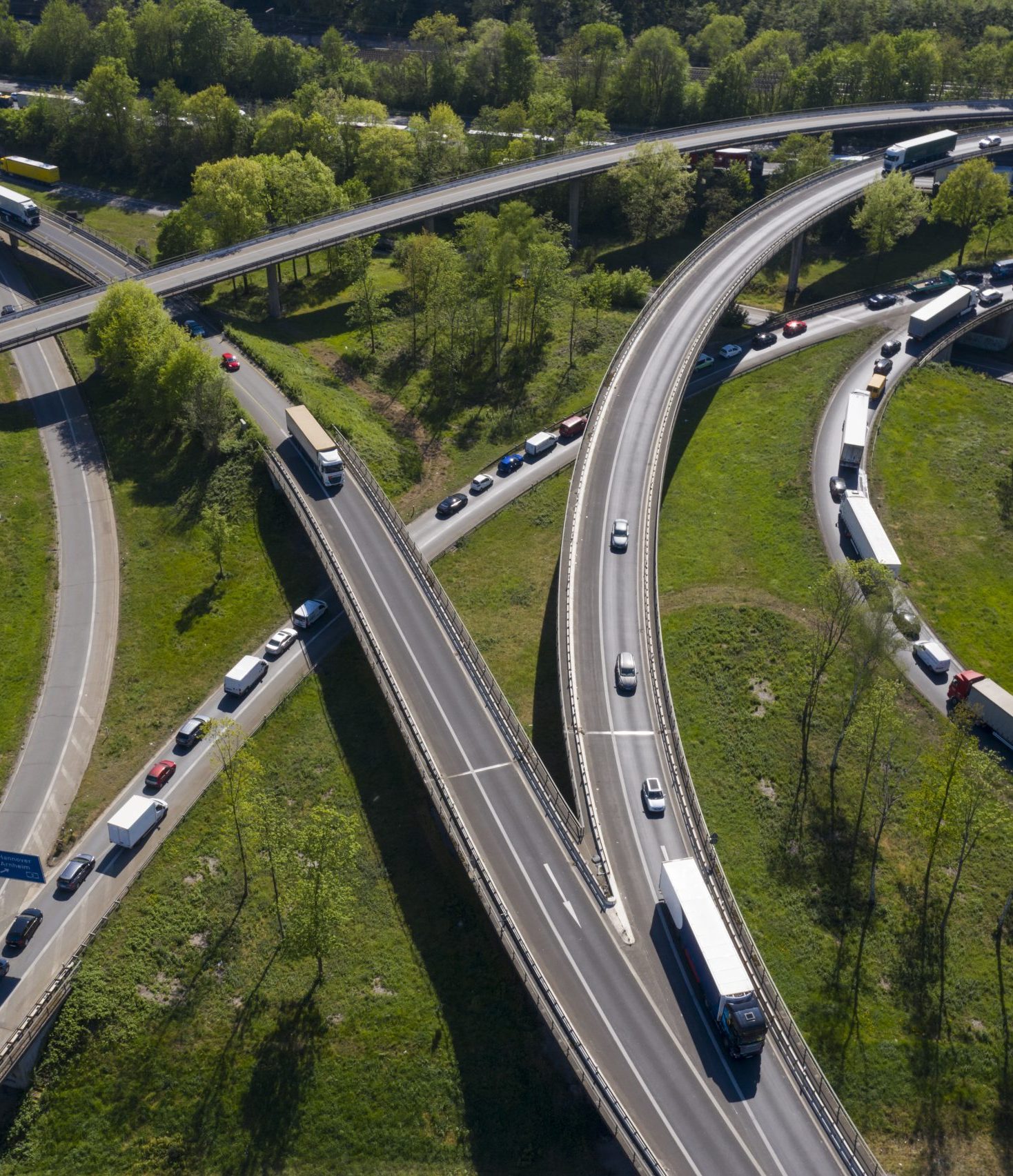 An aerial view of a highway interchange with multiple elevated roadways curving over a green landscape. Traffic is visible on all roads, with a mix of cars and trucks moving or waiting, particularly on ramps leading to or from the highways. The surrounding area is lush with trees and grass, creating a contrast between the natural environment and the concrete infrastructure.