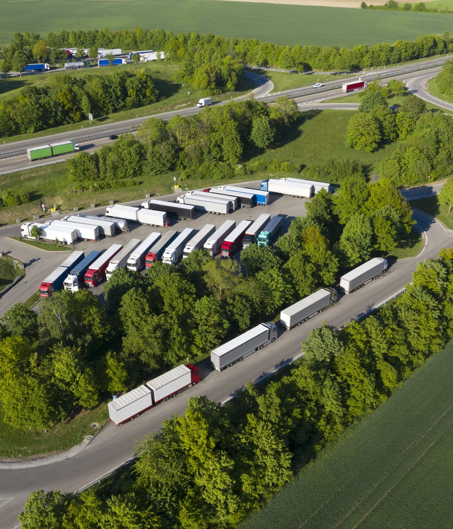 An aerial view of a rest area for trucks located near a highway, surrounded by lush green trees and fields. Multiple semi-trucks with trailers are parked in organized rows within the designated parking area, while others wait along a nearby road. The scene includes a highway in the background with vehicles in motion, connecting to curved access roads leading to the rest stop.