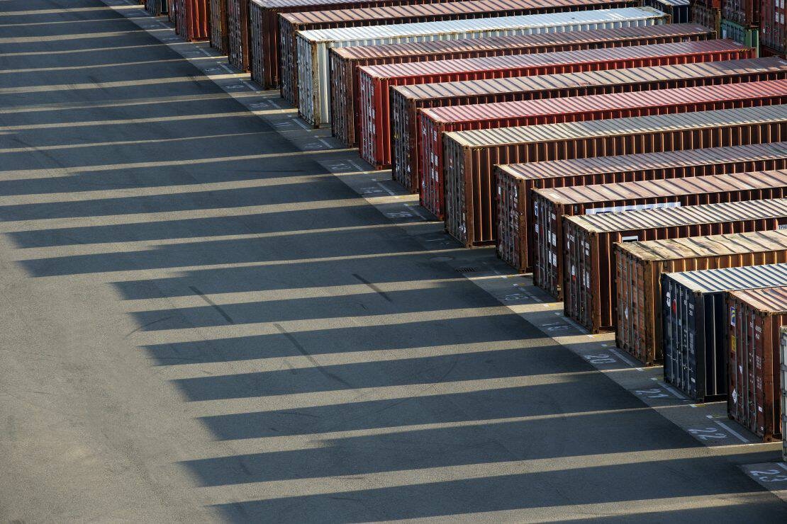 Rows of shipping containers neatly aligned in a logistics or storage yard, casting long, sharp shadows on the asphalt from the bright sunlight. The containers are a mix of colors, including red, brown, white, and black, with visible wear from use.