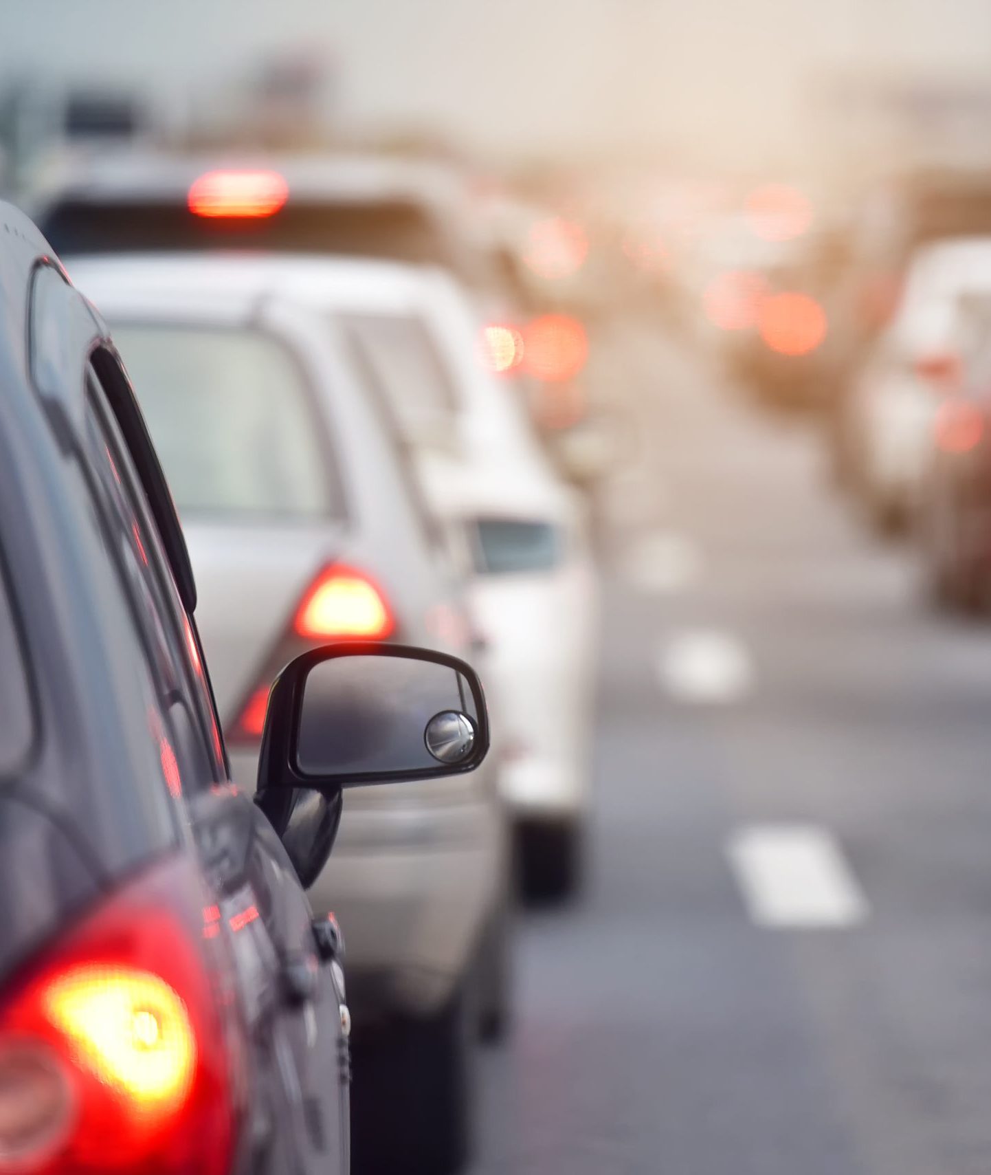 A close-up view of cars in a traffic jam on a multi-lane road. The image focuses on the side mirror of a dark-colored car in the foreground, with a line of vehicles stretching into the distance. Red brake lights and blurred bokeh effects emphasize the congestion during what appears to be daytime with soft natural light.